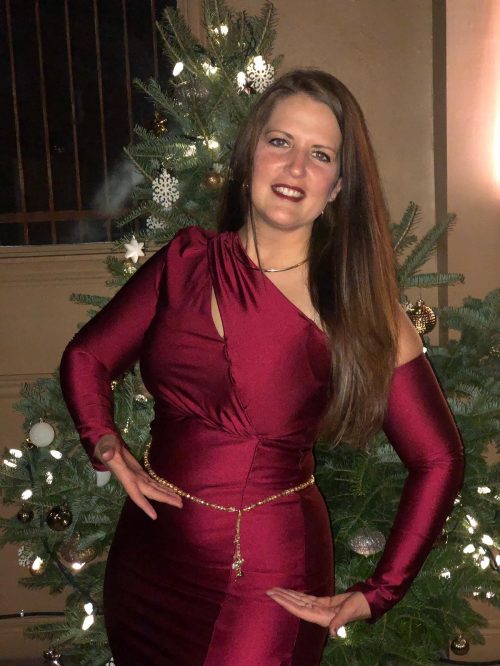 Customer posing in a satin maroon outfit wearing a golden bellybelt in front of a holiday tree.