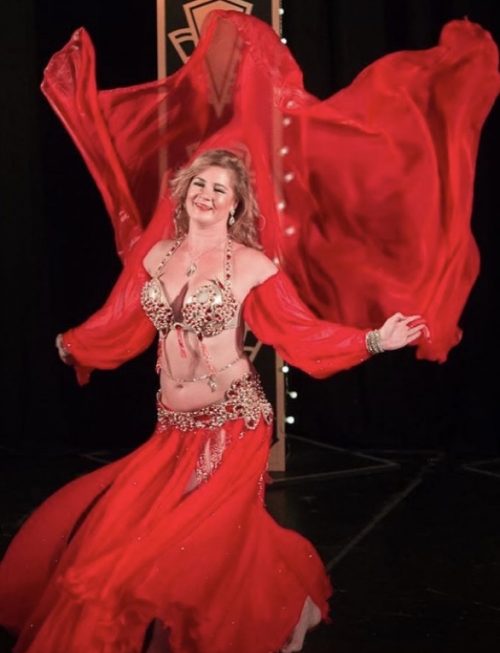 Bellydancer posing in a red outfit with a red fabric blowing in the wind wearing a clear and golden bellybelt.