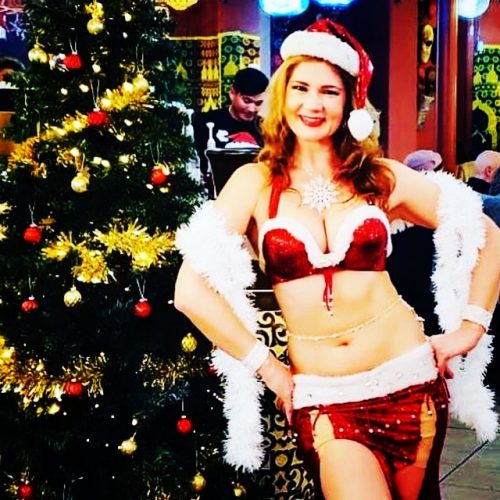 Customer smiling wearing a Santa hat and red and white bellydance outfit next to a holiday tree, wearing a bellybelt.